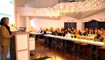 Sitzungssaal mit Teilnehmenden der Tagung und am Podium steht eine Frau und begrüßt